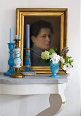 Blue candles in candlesticks and vase of flowers in front of oil painting on wall bracket