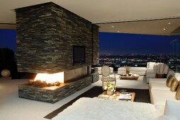 White sofa combination and open fireplace in stone-tiled partition; panoramic view of city by night in background