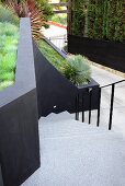 View down flight of pale concrete steps between black metal balustrade and black wall of terraced garden