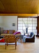 Wooden chair and armchair next to comfortable sofa with cushions in front of sliding wall element covered in Japanese paper in modern interior with wood-clad, barrel-vaulted ceiling