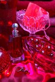 Still-life arrangement in red - perfume bottle next to various glass dishes on red satin cloth