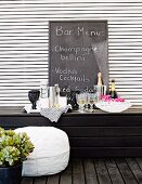 Improvised bar with glasses and bottles on dark wooden bench, menu written on chalkboard leaning on white slatted wall