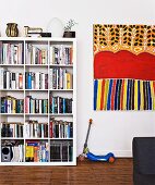 Weisses Bücherregal mit quadratischen Einteilungen, daneben Kinderroller und modernes, buntes Bild an Wand (Kunst der Aborigines)