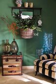 Sessel mit gestreiftem Bezug, Glasgefässe mit Lichterkette, Beistelltisch mit Ballonflaschen und Korb mit Weihnachtsdeko vor grüner Wand