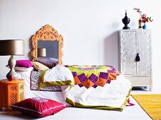 Bett mit gesteppter Tagesdecke und mehreren Kissen dekoriert, kunsthandwerklicher Spiegelrahmen und silberfarbener Schrank im Ethnostil