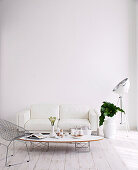 Wire mesh chair and white upholstered sofa around coffee table, photo lamp on the side and floor vase with green leaves