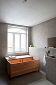 Designer wooden bathtub with open lid below restored window, concrete washstand and concrete wall facing