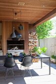 Black shell chairs with delicate metal frames at long table on roofed terrace with fireplace and firewood stacked against wooden wall in courtyard