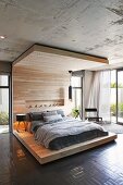 Comfortable double bed on modern, floor-to-ceiling bed platform with canopy in light-flooded interior with concrete ceiling and open terrace doors