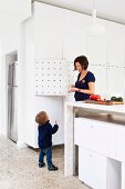 Mother and child in open-plan kitchen; stone counter with white modules below and floor-to-ceiling white fitted cupboards