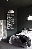 Black bedroom with white accents; French bed below porthole window, white pendant lamp, white screen and white lettering on black panel on wall