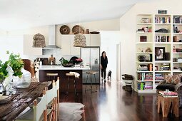Shelving, dining table with rustic wooden top, woman and dog in background and wicker lamps in kitchen in modern, open-plan interior
