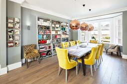 Room with bay window in English house; long table, yellow upholstered chairs, board floor and grey bookcases