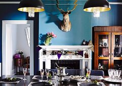 Dining room in eclectic mixture of styles with festively set table, antique glass-fronted cabinet & hunting trophy above open fireplace