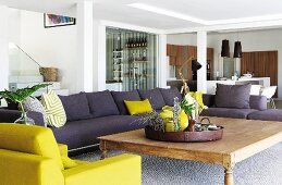 Purple-grey sofa and yellow armchair around rustic wooden coffee table in spacious living room with dining area behind pillars