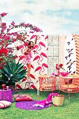 Different flamingo print patterns, outdoor furniture and accessories in shades of red and purple; woman standing on stool hanging up fairy lights