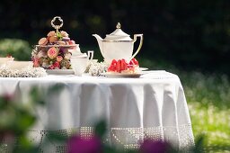 Garden table romantically set with white tablecloth, gold-rimmed tea service and opulent cake stand decorated with flowers