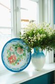 Floral decorative plate next to bouquet of chamomile in pale blue jug on sill of lattice window