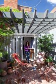 Deckchairs on terrace below grey-painted wooden lattice pergola; woman walking out of house