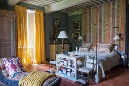 Chaise longue opposite chairs and half-height shelves at foot of bed; floor-length, yellow curtains at window of country-style bedroom