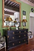 Table lamps with gold lampshades on black-stained chest of drawers below mirror on green wall