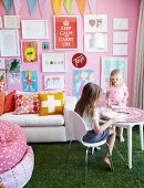 Children playing at round table, white sofa below collection of framed children's drawings on pink wall and green, lawn-effect carpet