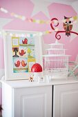 Framed comic page and birdcage on white cabinet against pink mural with stylised owl