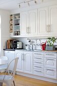 White fitted kitchen with panel doors and strip handles; dining area with vintage chairs