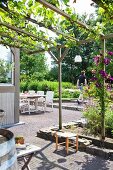 Gepflasterte Terrasse mit berankter Pergola, im Hintergrund ein gedeckter Tisch und Garten