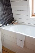 Detail of white, wood-clad bathtub with white clapboard side wall and dark, mosaic-tiled back wall