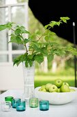 Vase of leafy twigs and bowl of green apples behind tealight holders on garden table