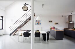 Minimalistische Loftwohnung mit Retro Flair, weisser Esstisch und schwarze Stühle vor Treppenaufgang, seitlich offene Küche mit dunkelgrauen Schränken