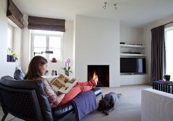 Woman sitting on chaise longue and dog on floor in cosy atmosphere in front of open fire in modern interior