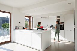White, minimalist, designer kitchen; small child sitting on kitchen counter and mother opening wall cupboard