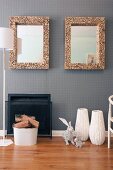Hare ornaments in front of white floor vases next to open fireplace and bucket of logs; two framed mirrors on wall with grey wallpaper