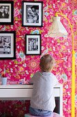 Child sitting at desk, standard lamp with nostalgic fabric lampshade and yellow-painted base, family photos on pink wallpaper with floral pattern