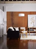 Wohnzimmer mit Flokati und schwarzem Sofa, arrangiert mit gerahmtem Goldbuchstaben-Bild vor Wandgestaltung in Holzoptik