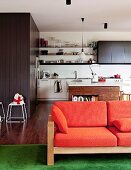 Sofa with wooden frame and orange upholstery in front of open-plan kitchen; appliances built into grey wood panelling and island counter with bookshelf