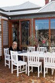 Frau auf Terrasse, weisslackierte Outdoorstühle und Tisch auf Ziegelboden vor Holzhaus mit raumhohen Terrassenfenstern
