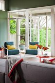Sofa opposite blue easy chairs with yellow scatter cushions next to open terrace doors with view into garden