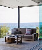 Coffee table and matching sofa with brown cushions and patterned scatter cushions against glass balustrade in relaxation area on roofed wooden deck with view of Pacific Ocean