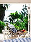 Bamboo sun lounger and white wicker table on sunny terrace with sea view and tropical plants