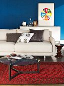 Simple, beige sofa and glass table on red, patterned Oriental rug; picture of colourful wheel on blue-painted wall