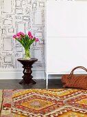 Vase of tulips and designer stool against wall with pattern of rectangles; kilim rug with traditional pattern in foreground