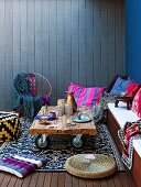 Upholstered bench with colourful scatter cushions and rustic side table on castors against blue, wood cladding