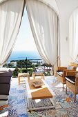 Sitzmöbel in Loggia mit bunten, mediterranen Bodenfliesen; Blick durch einen Rundbogen mit drapierten Vorhängen auf das Meer vor Capri