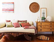 Tagesbett mit hellen Polstern und farbigen Kissen auf Holzgestell neben Kommode vor weisser Holzwand