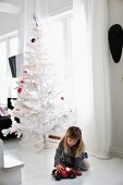 Girl playing with toy car on white wooden floor in front of white, artificial Christmas tree with purple baubles