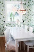 Dining table and chairs painted white below traditional chandelier with glass pendants in rustic dining area with pastel floral wallpaper and festive star ornaments in windows