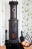 Vintage, cylindrical wood-burning stove and basket of firewood in corner of room with romantic floral wallpaper